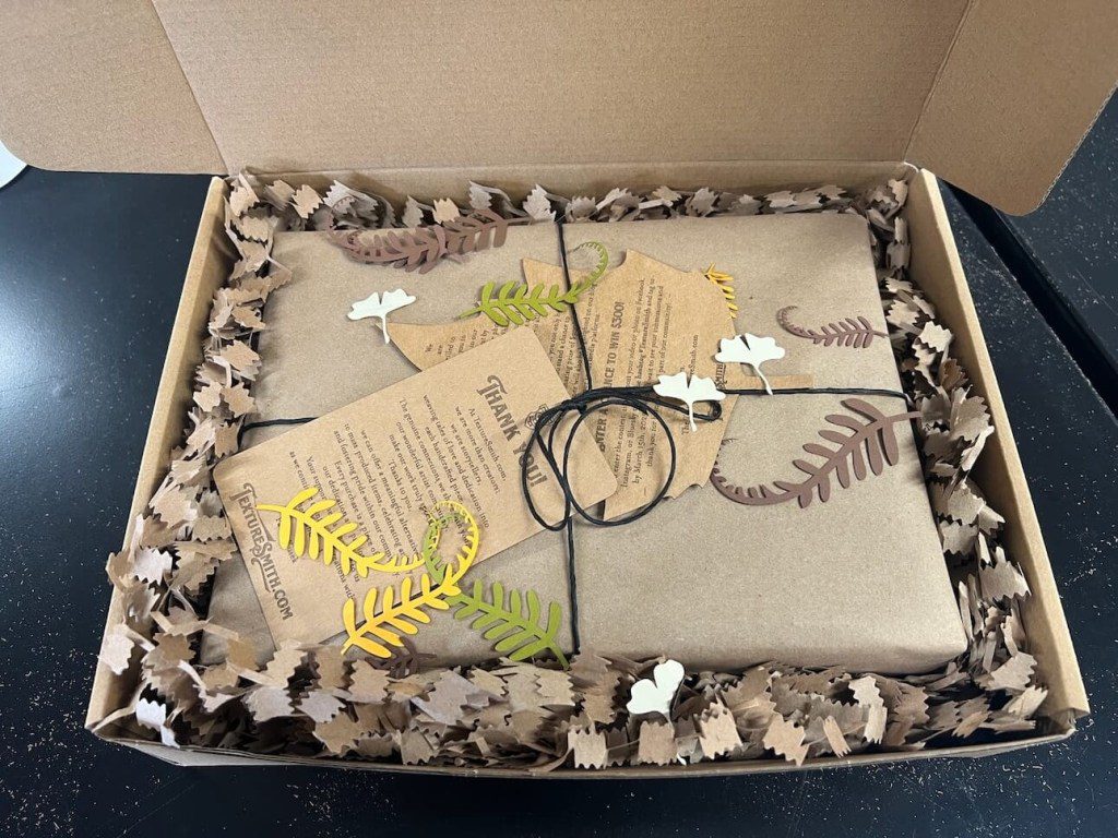 Gift box with wrapped present, fern leaves, and tags.
