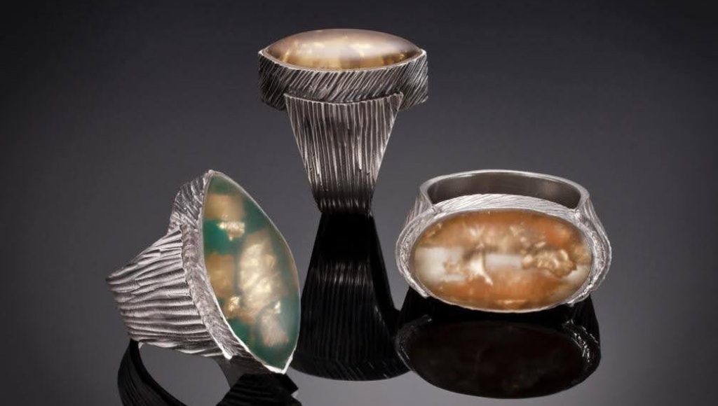 Three unique silver and stone decorative vessels on a dark reflective surface.