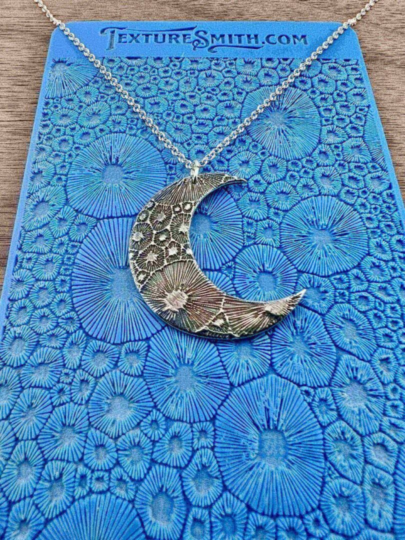 Silver crescent moon pendant with floral details on blue textured surface.