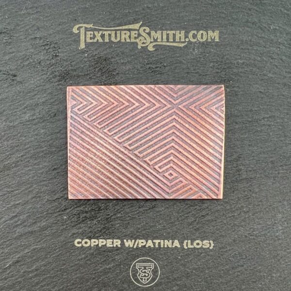 Rectangular copper sheet with a geometric patina pattern on a dark surface.