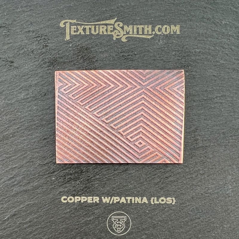Rectangular copper sheet with a geometric patina pattern on a dark surface.