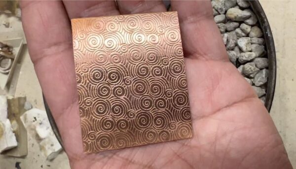 Hand holding a copper sheet with intricate spiral patterns.