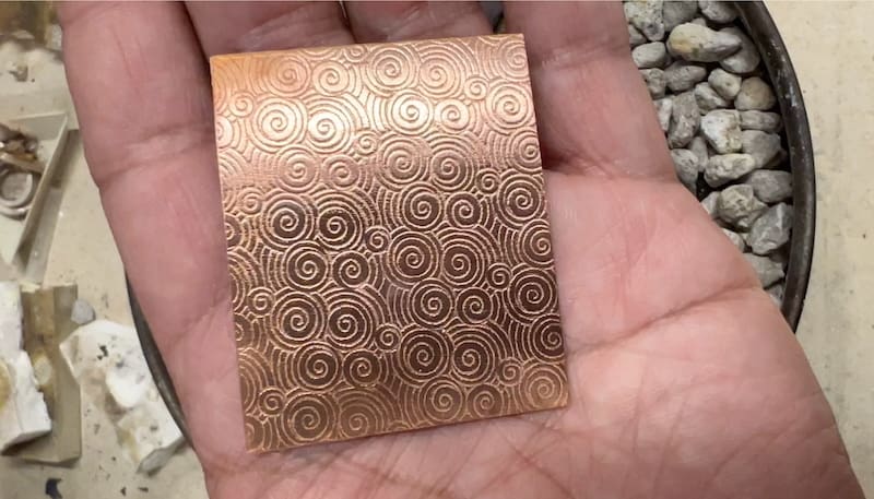 Hand holding a copper sheet with intricate spiral patterns.