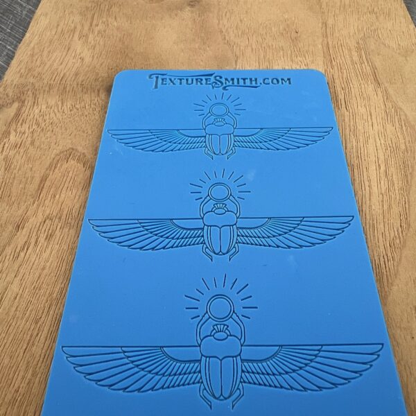A blue stencil with three Egyptian scarab wing designs on a wooden surface.